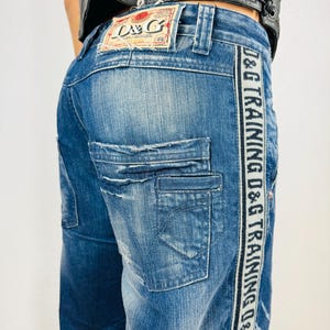 Puede incluir: Vaqueros de mezclilla azul con un aspecto desgastado y una raya D&G Training en blanco y negro que recorre la costura lateral. Los vaqueros tienen un bolsillo trasero con una etiqueta D&G.