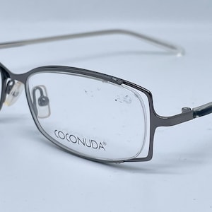Könnte beinhalten: Eine Brille aus schwarzem Metall mit klaren Gläsern. Der Markenname "COCONUDA" ist auf dem Rahmen aufgedruckt.