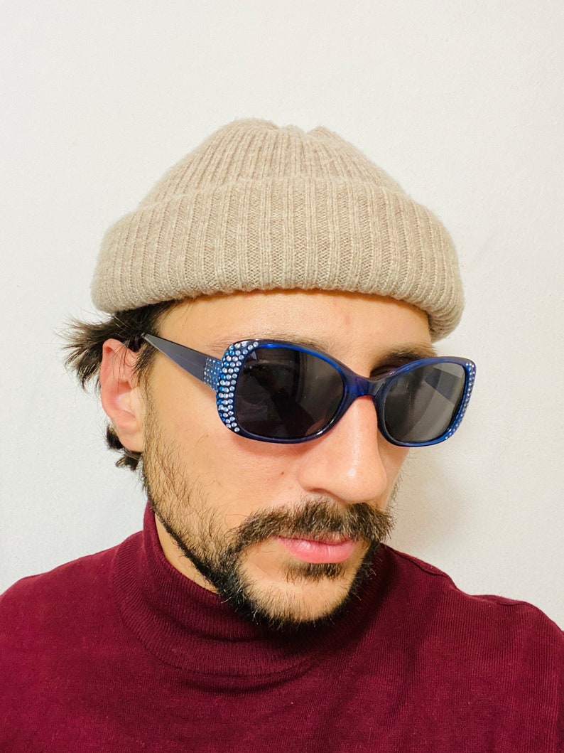 Puede incluir: Una persona con una boina beige y gafas de sol azules con pedrer&iacute;a. Las gafas tienen lentes oscuras. La persona lleva un jersey de cuello alto marr&oacute;n.