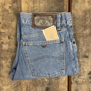 May include: Light blue denim jeans featuring a brown leather patch on the waistband with the text Armani Jeans Established 1981 Made in Italy. A small metal eagle logo is centered on the back pocket, which also contains a beige paper tag.