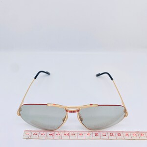 May include: Gold-tone aviator sunglasses with gray lenses and red accents. The sunglasses are measured with a ruler, showing a width of approximately 5 inches.