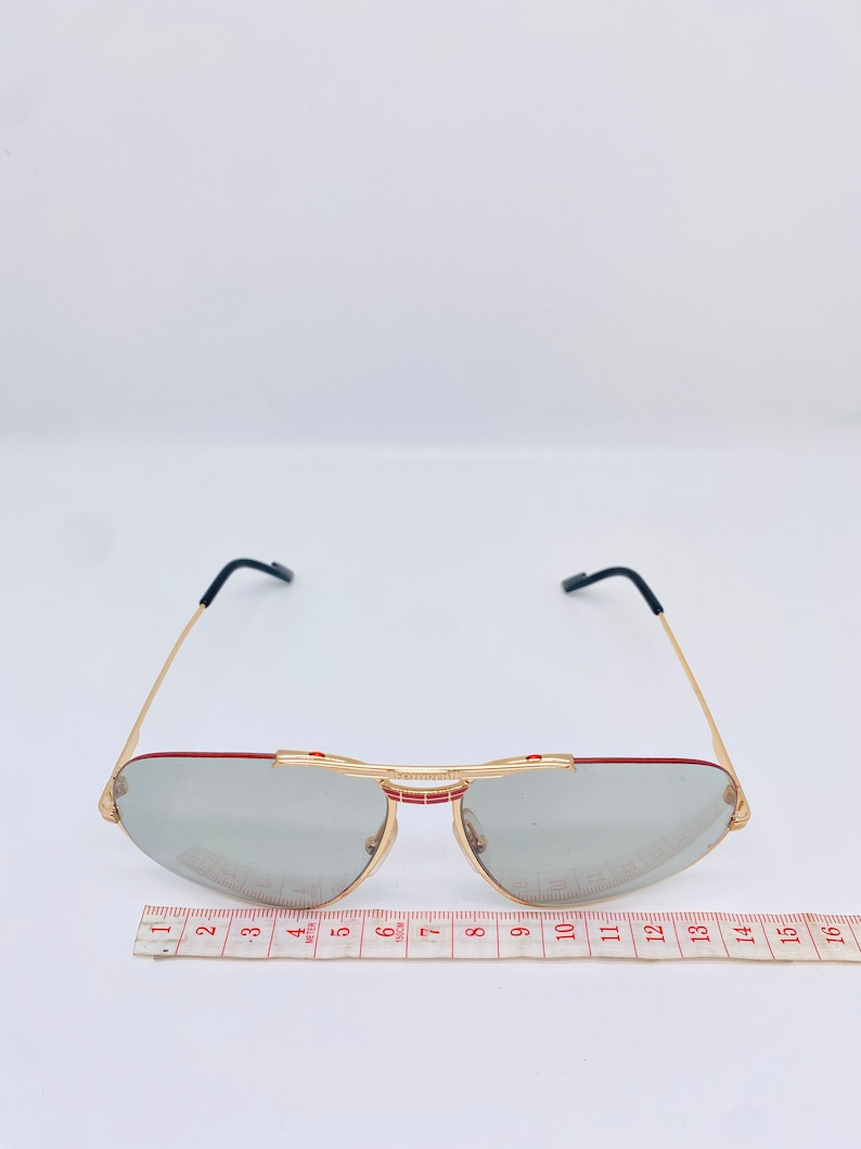 May include: Gold-tone aviator sunglasses with gray lenses and red accents. The sunglasses are measured with a ruler, showing a width of approximately 5 inches.