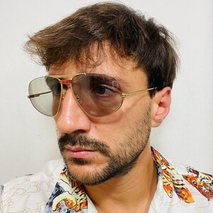 May include: A man wearing gold aviator sunglasses with brown lenses and a white shirt with a tiger print.