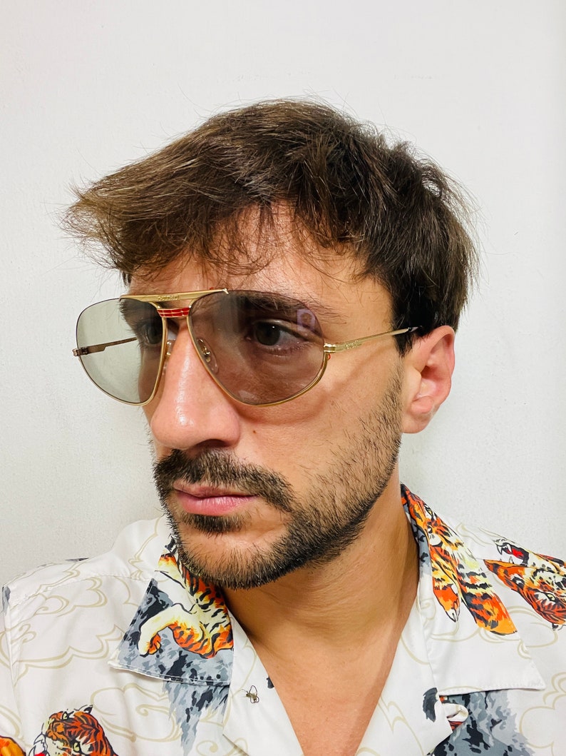 May include: A man wearing gold aviator sunglasses with brown lenses and a white shirt with a tiger print.