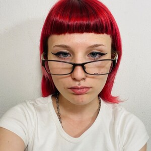 May include: A person with bright red hair and black-framed glasses. They are wearing a white t-shirt with the text "Radio Silence" in a black box. A silver chain necklace and facial piercings are also visible.