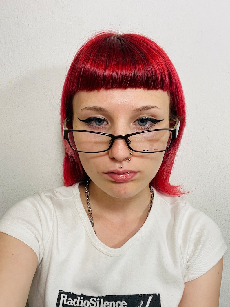 May include: A person with bright red hair and black-framed glasses. They are wearing a white t-shirt with the text "Radio Silence" in a black box. A silver chain necklace and facial piercings are also visible.