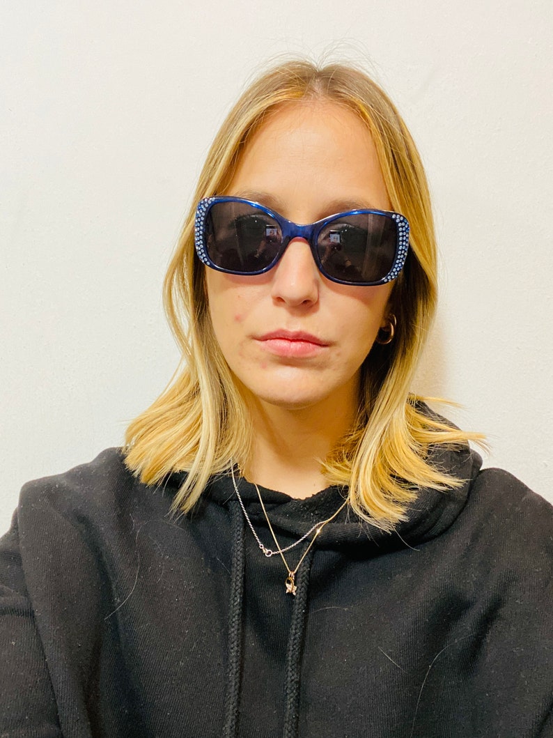 Puede incluir: Una mujer con una sudadera con capucha negra, un collar de oro y gafas de sol azules con pedrer&iacute;a.