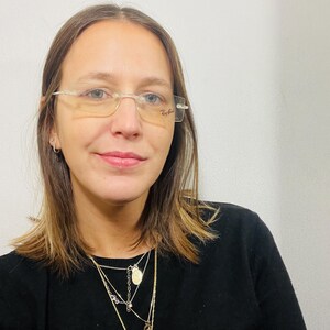 May include: A woman with brown hair wearing clear framed glasses and a black sweater with a layered gold necklace.