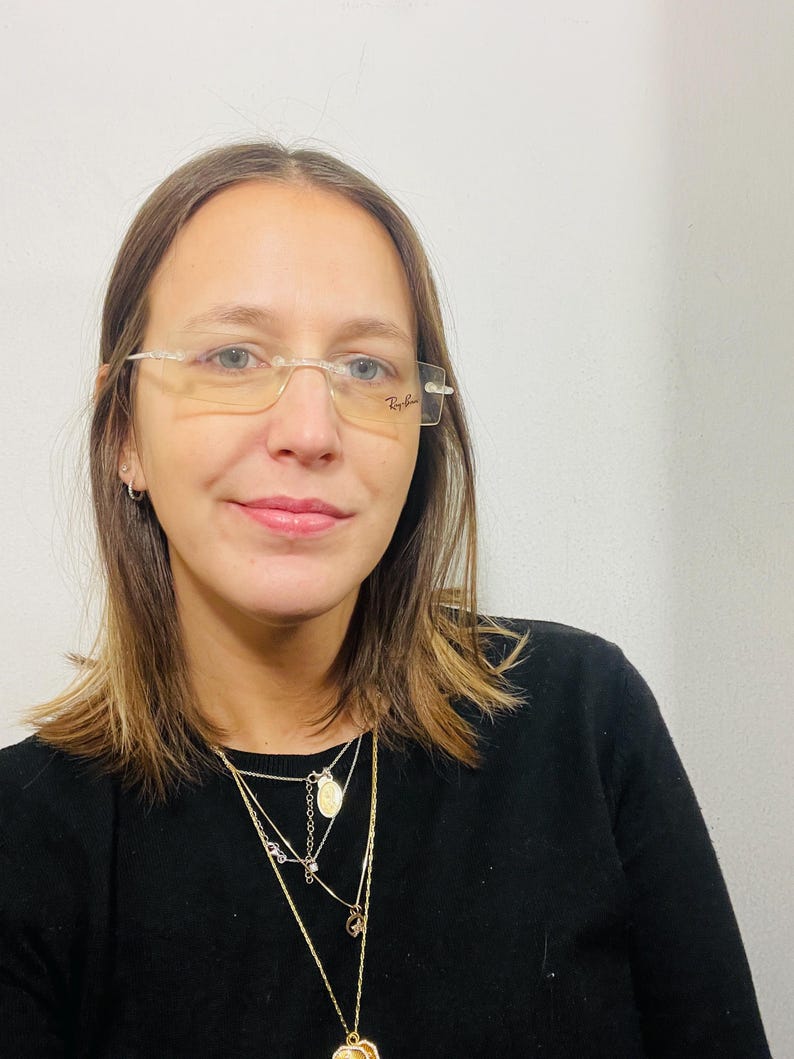 May include: A woman with brown hair wearing clear framed glasses and a black sweater with a layered gold necklace.