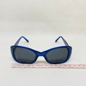 Puede incluir: Gafas de sol de pl&aacute;stico azul con pedrer&iacute;a alrededor de las monturas. Las gafas tienen lentes oscuras y se miden en una regla.
