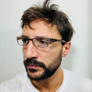 Może przedstawiać: Mężczyzna z brązowymi włosami i brodą nosi okulary w brązowej oprawce. Ma na sobie biały t-shirt.