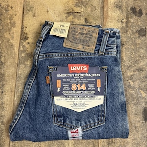 Può includere: Un paio di jeans in denim blu a lavaggio medio con una toppa in similpelle marrone sulla cintura e una grande etichetta di cartone sulla tasca posteriore. L'etichetta riporta il testo Levi's America's Original Jeans 814 Genuine Quality Clothing.