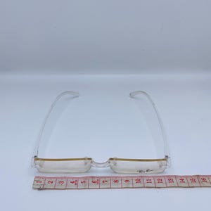 May include: Clear plastic frame glasses with a gold trim and the text "Ray-Ban" on the right lens. The glasses are measured on a ruler that shows they are 12 inches wide.