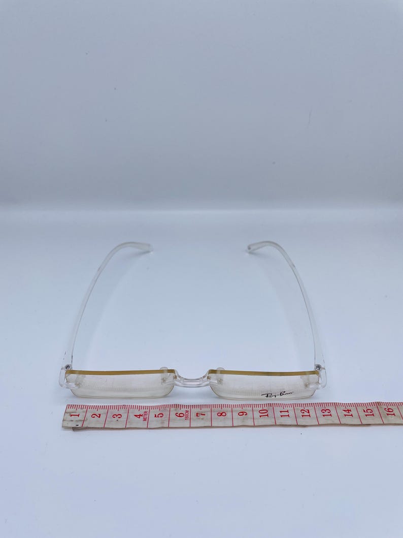May include: Clear plastic frame glasses with a gold trim and the text "Ray-Ban" on the right lens. The glasses are measured on a ruler that shows they are 12 inches wide.