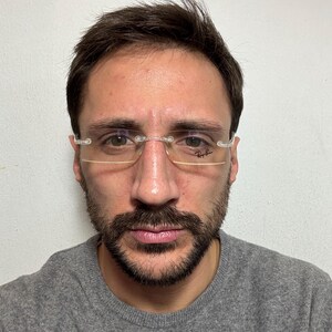 May include: A man with short brown hair and a beard is wearing clear-framed glasses with the Ray-Ban logo on the right lens. He is wearing a gray sweater.