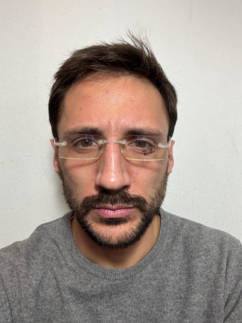 May include: A man with short brown hair and a beard is wearing clear-framed glasses with the Ray-Ban logo on the right lens. He is wearing a gray sweater.