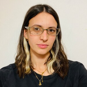 Może przedstawiać: Osoba z długimi brązowymi i blond włosami w czarnym t-shircie i złotych naszyjnikach z małym wisiorem w kształcie ośmiornicy. Nosi okulary i ma kolczyk w nosie.
