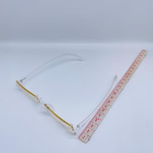 May include: Clear plastic rectangular frame glasses with a gold trim. The glasses are lying on a white surface next to a measuring tape.