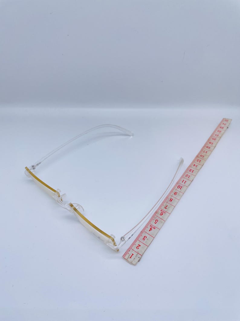 May include: Clear plastic rectangular frame glasses with a gold trim. The glasses are lying on a white surface next to a measuring tape.
