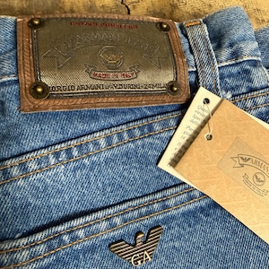 Puede incluir: Primer plano de unos vaqueros de mezclilla azules con un parche de cuero marrón y un emblema metálico. El parche dice "ARMANI JEANS" y "MADE IN ITALY". Una etiqueta de papel con el logo de Armani está adjunta.