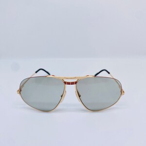 May include: Gold aviator sunglasses with gray lenses and a red and black stripe detail on the frame.