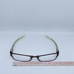 May include: A pair of eyeglasses with a dark brown frame and clear lenses. The temples are a light green color. A measuring tape is placed below the glasses, showing a length of approximately 6 inches.