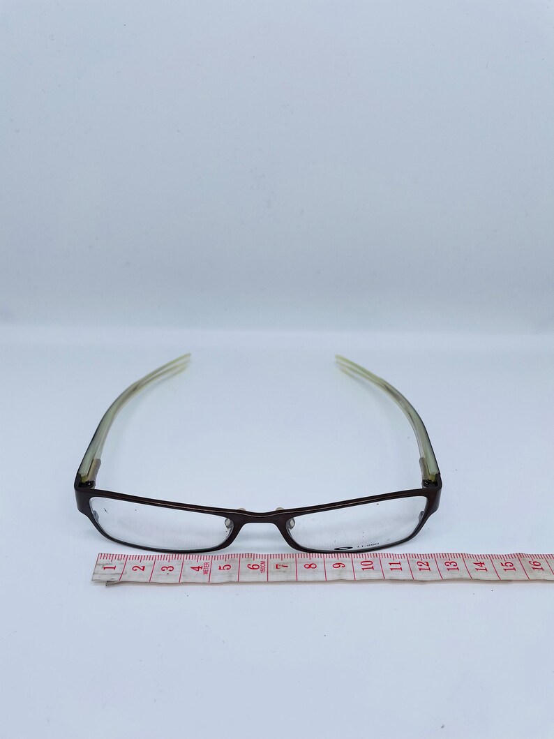 May include: A pair of eyeglasses with a dark brown frame and clear lenses. The temples are a light green color. A measuring tape is placed below the glasses, showing a length of approximately 6 inches.