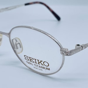 Könnte beinhalten: Silberfarbene Metallbrille mit rundem Gestell und einem dekorativen Design an den Bügeln. Die Brille hat klare Gläser mit dem Text "SEIKO 100% TITANIUM DEMO LENS" auf dem rechten Glas.