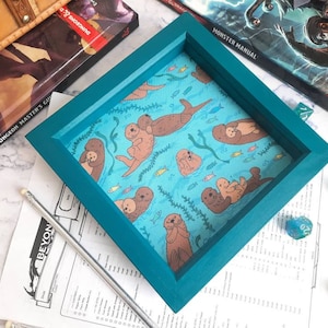 May include: Teal blue wooden dice tray with a light blue fabric insert featuring a repeating pattern of sea otters and fish.