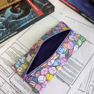 May include: A purple zippered pencil case with a colorful print of dice and gemstones. The case is open and the inside is lined with purple fabric.