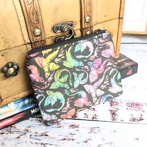 May include: A black zippered pouch with a colorful watercolor dragon print. The pouch is lying on a wooden surface.