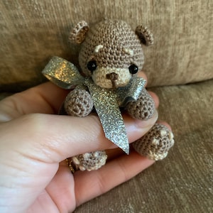 May include: A small brown crocheted teddy bear wearing a silver glitter bow. The bear is sitting in a hand.