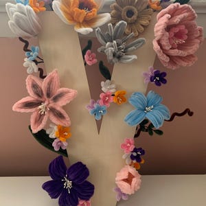 May include: A wooden letter "V" adorned with an assortment of colourful felt flowers. The flowers feature shades of pink, orange, blue, purple, and white. The flowers are arranged around the letter, creating a floral design. The background is a light pink.