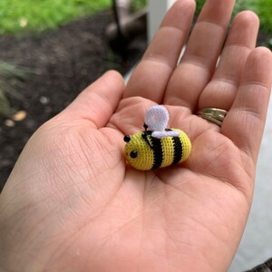 May include: A small crocheted bee with yellow and black stripes, white wings, and black eyes. The bee is sitting in the palm of a hand.