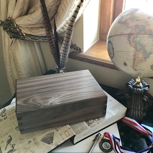 May include: A dark wood box with a hinged lid, resting on a book featuring a vintage map design. The box has a natural wood grain pattern and is closed. The book is on a table with a globe and other decorative items.
