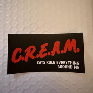 May include: A black rectangular sticker with the word "C.R.E.A.M." in red, stylized lettering. Below the acronym, the text reads "CATS RULE EVERYTHING AROUND ME" in white. The sticker is on a white, textured background.