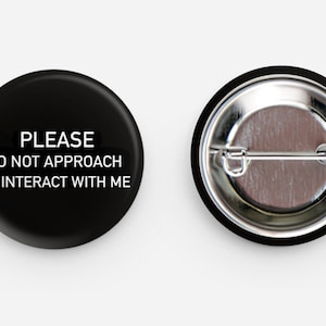Op de afbeelding: Een zwarte knop met witte tekst die "PLEASE DO NOT APPROACH OR INTERACT WITH ME" luidt. De achterkant van de knop is zilverkleurig met een pin.