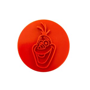May include: A bright orange, circular stamp featuring a smiling cartoon face with a carrot nose and twig-like hair. The stamp is a relief design, with the character's features raised from the surface. The design is centered on the stamp.
