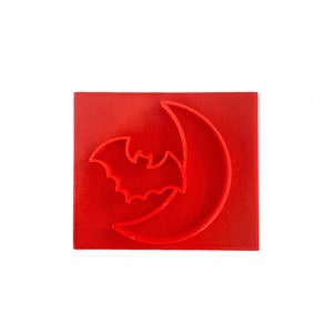 May include: A red, square-shaped cookie cutter featuring a bat silhouette in front of a crescent moon. The design is a Halloween theme, perfect for baking spooky treats. The cutter is made of a solid, textured material.