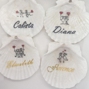 May include: Four white scallop shells, each inscribed with a name and a small drawing. The names are Calista, Diana, Elizabeth, and Florence. The drawings show stick figures with hearts, with names in black or gold script.