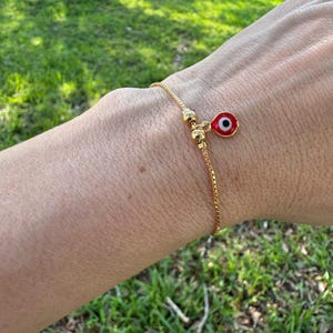 Op de afbeelding: Goudkleurige armband met een kleine, rood-witte evil eye bedel. De bedel is rond met een zwart centrum. De armband wordt om een pols gedragen, met een groene grasachtige achtergrond.