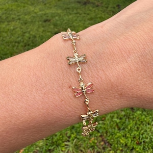 Peut inclure: Un bracelet doré orné de breloques libellules aux ailes roses et transparentes. Le bracelet est relié par de petites chaînes dorées et est porté sur un poignet, sur fond d'herbe verte et d'eau.