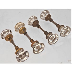 May include: Four antique glass door knobs with faceted crystal-like designs. The knobs are mounted on aged metal stems with brass-colored accents. The arrangement is on a white background, showcasing the vintage hardware.