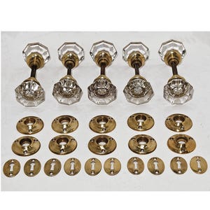 May include: Five antique door knobs with clear faceted glass and brass hardware. The set includes matching brass backplates and keyhole covers, perfect for restoration projects. The knobs have a classic, vintage aesthetic.