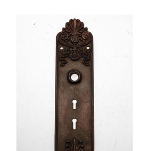 May include: A vintage, ornate, bronze door plate with a keyhole and a round knob. The plate has a decorative design with intricate details.