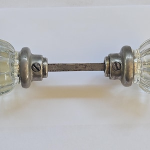May include: A vintage door knob set with clear, ribbed glass knobs and a metal rod. The knobs have a decorative, rounded shape and are attached to a metal base. The metal components show signs of age and wear.