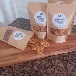 May include: Three brown paper bags filled with peanut brittle. Each bag has a white sticker with a moose and the text "Mamma Moose's Peanut Brittle".