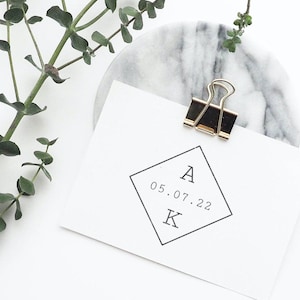 Pode incluir: Um cartão branco com as letras "A" e "K" e a data "05.07.22" impressas em forma de diamante, preso por um clip dourado e preto. O cartão repousa sobre um porta-copos de mármore, com raminhos verdes no fundo.