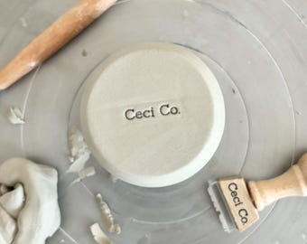 May include: A pottery wheel with tools and clay. A light gray ceramic piece with the text "Ceci Co." is in the center. A wooden handled tool and a clay stamp with the text "Ceci Co." are also visible.