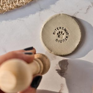 Puede incluir: Un disco circular de arcilla beige con una huella de pata estampada y el texto BITERSWIT STUDIO. Un sello con mango de madera se sitúa sobre la arcilla, y una herramienta de escultura de metal con mango de madera se encuentra a la derecha.